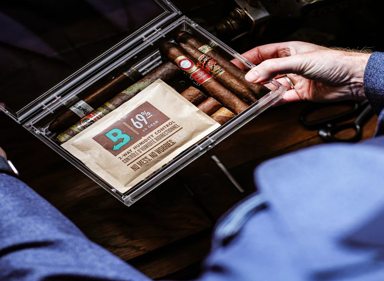 Wooden box of cigars on a table, surrounded by loose tobacco, bottles, and a potted plant; warm lighting